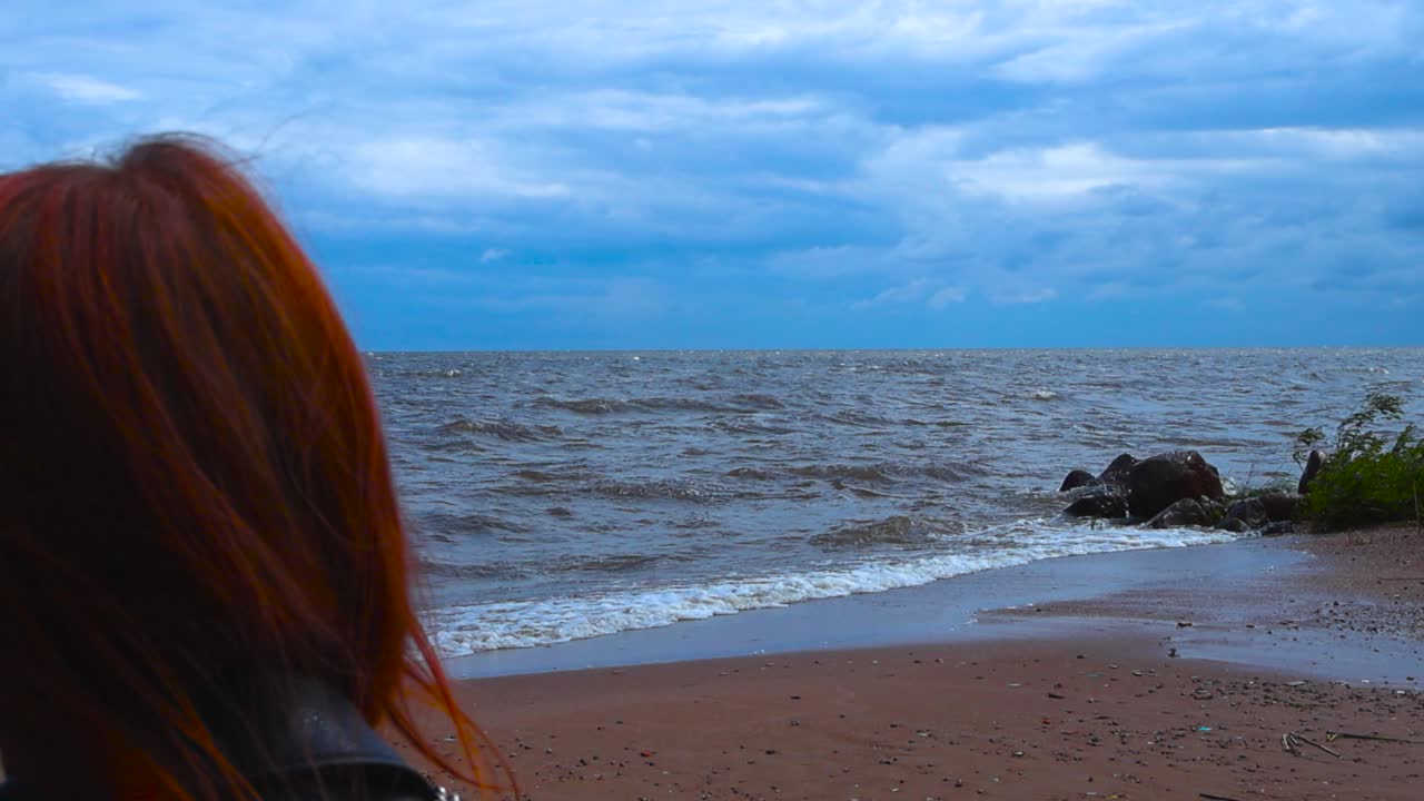 Red haired woman watching foamy and bubbly white water Peipsi lake water waves crashing and hitting a sandy red shoreline during a windy summer day with blue sky that have white clouds. Plants move