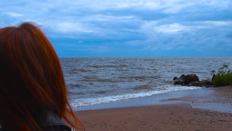 red haired woman watching foamy and bubbly white water peipsi lake water waves crashing and hitting a sandy red shoreline during a windy summer day with blue sky that have white clouds. plants move