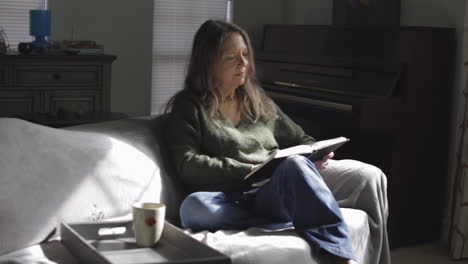 attractive adult woman reading book on the sofa with coffee mug on her side