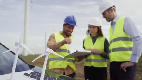 Three-caucasian-and-latin-engineers-standing-on-wind-turbine-field-and-discussing-over-plastic-models.