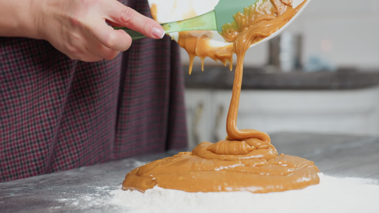 녹색 스파라를 사용하여 카운터에 두꺼운 카라멜 색의 반죽을 밀가루로 부는 여성의 클로즈업, 부드럽고 풍부한 혼합물이  그에서 흐릅니다.