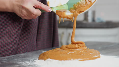 close-up of woman pouring thick caramel-colored batter into flour on countertop using green spatula, smooth, rich mixture flows from white bowl