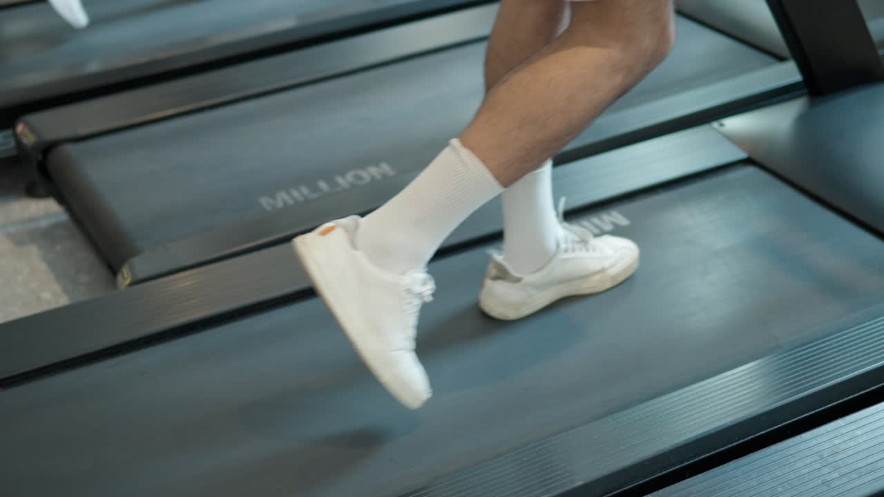 Legs of a man in white sport shoes walking on treadmill at indoor gym - slowmo