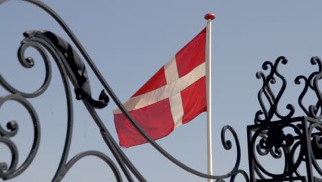 Alter-Zaun-Und-Wehende-Dänische-Flagge-Vor-Blauem-Himmel-Mit-Einem-Schwan,-Der-Durch-Das-Bild-Zieht