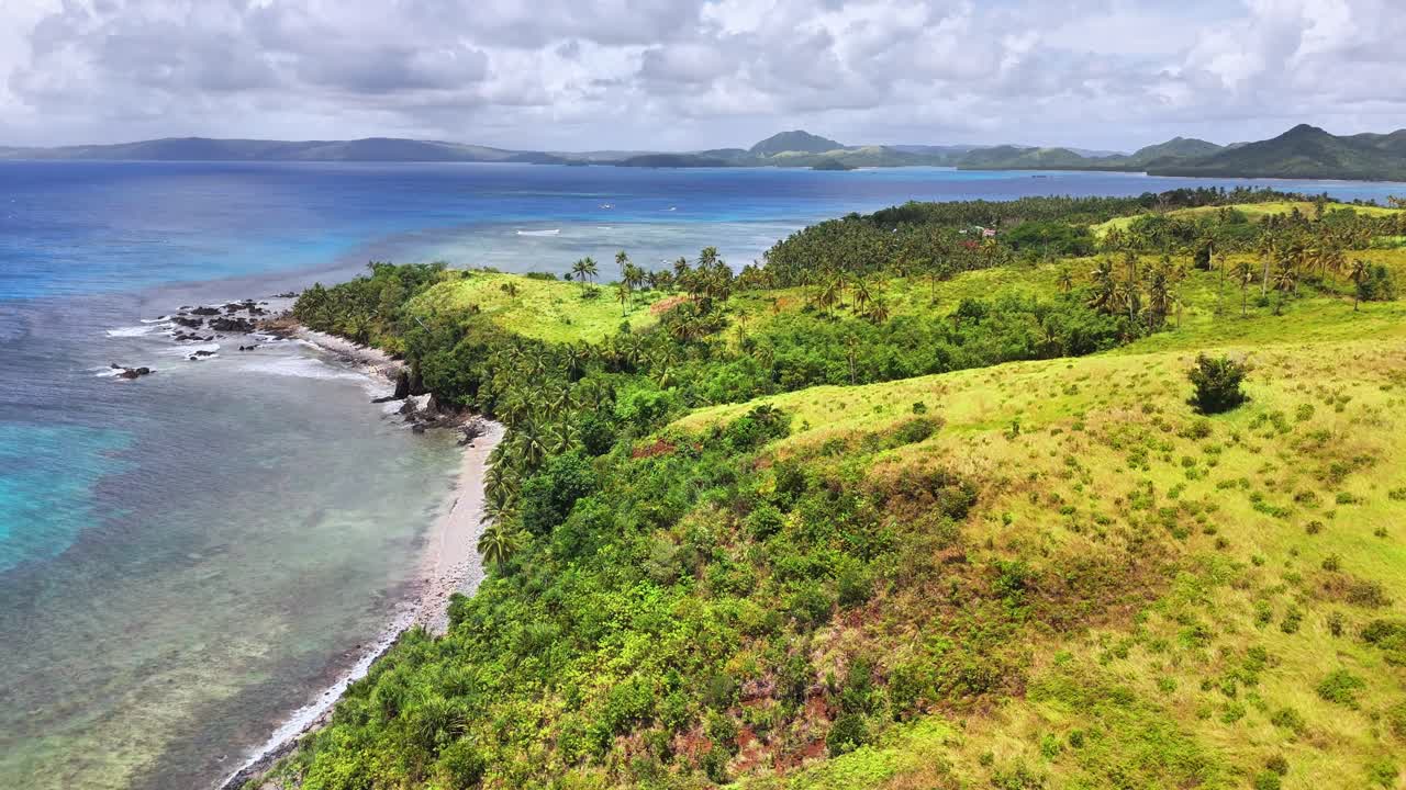 Lush green hills meet turquoise waters along the tropical coastline of Corregidor Island, Siargao, Philippines, featuring palm trees, rocky shores, and distant mountainous islets under cloudy skies