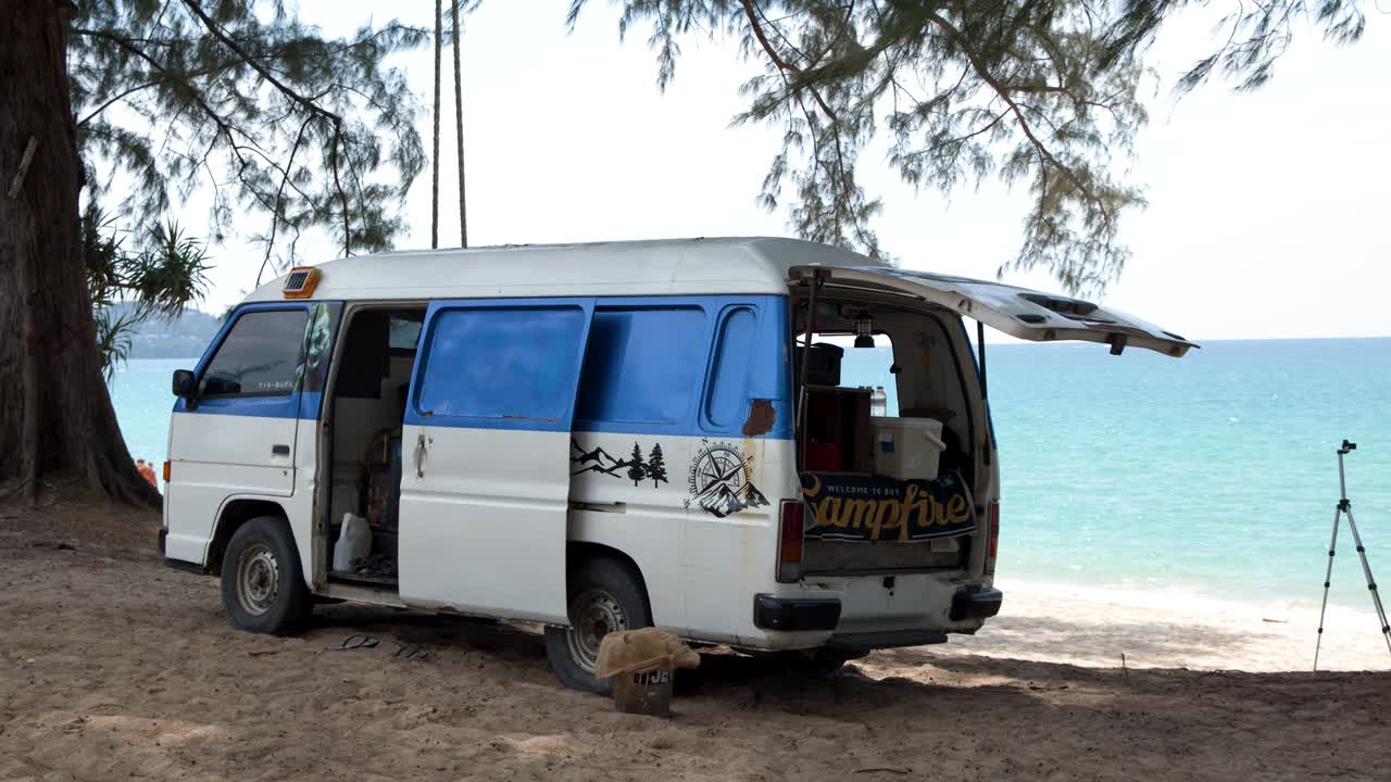 A stationary wide shot of a camper van parked under trees by the turquoise ocean