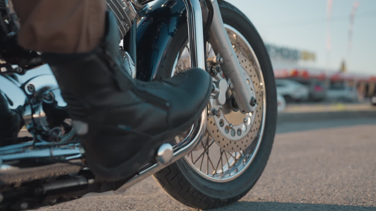 Lower angle close up of motorcycle front wheel and brake disc with rider placing leg on bike stand, chrome engine and metal parts reflecting light, urban street background