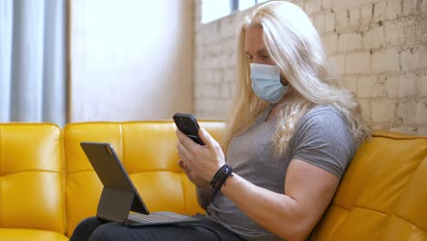 masked long blonde hair bearded man on yellow vegan leather vinyl couch typing on ipad pro magic keyboard then picking up iphone 13 pro max and texting messaging back - in cinema 4k