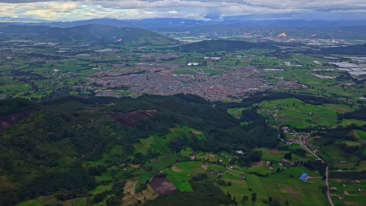 Drone approaches Zipaquirá from above, flying over green valley slopes and farmland toward the city in the distance under a cloudy sky