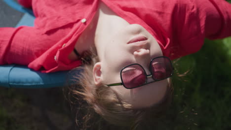 elegant woman wearing sunglasses lies back on swing with relaxed expression, soaking up warm sun, close up captures peaceful moment as she gently swings in grassy backyard, eyes closed in total calm