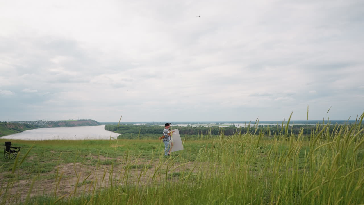 먼 마을을 향해 강을 바라보는 잔디 언덕에 있는 한 부부, 카레드 셔츠와 모자를 입은 남자,  옷을 입은 여자, 앞면에 검은 의자가 있는 높은 초록 잔디를 고 고 있다.