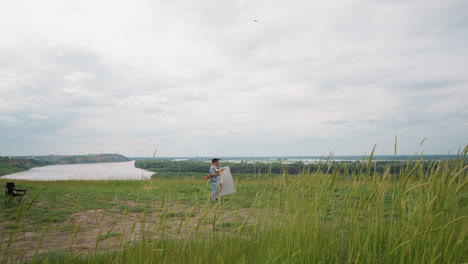 a couple on a grassy hill, looking out over a river toward a distant town. the man in a plaid shirt and hat, and the woman in white, walk through tall green grass with a black chair in the foreground