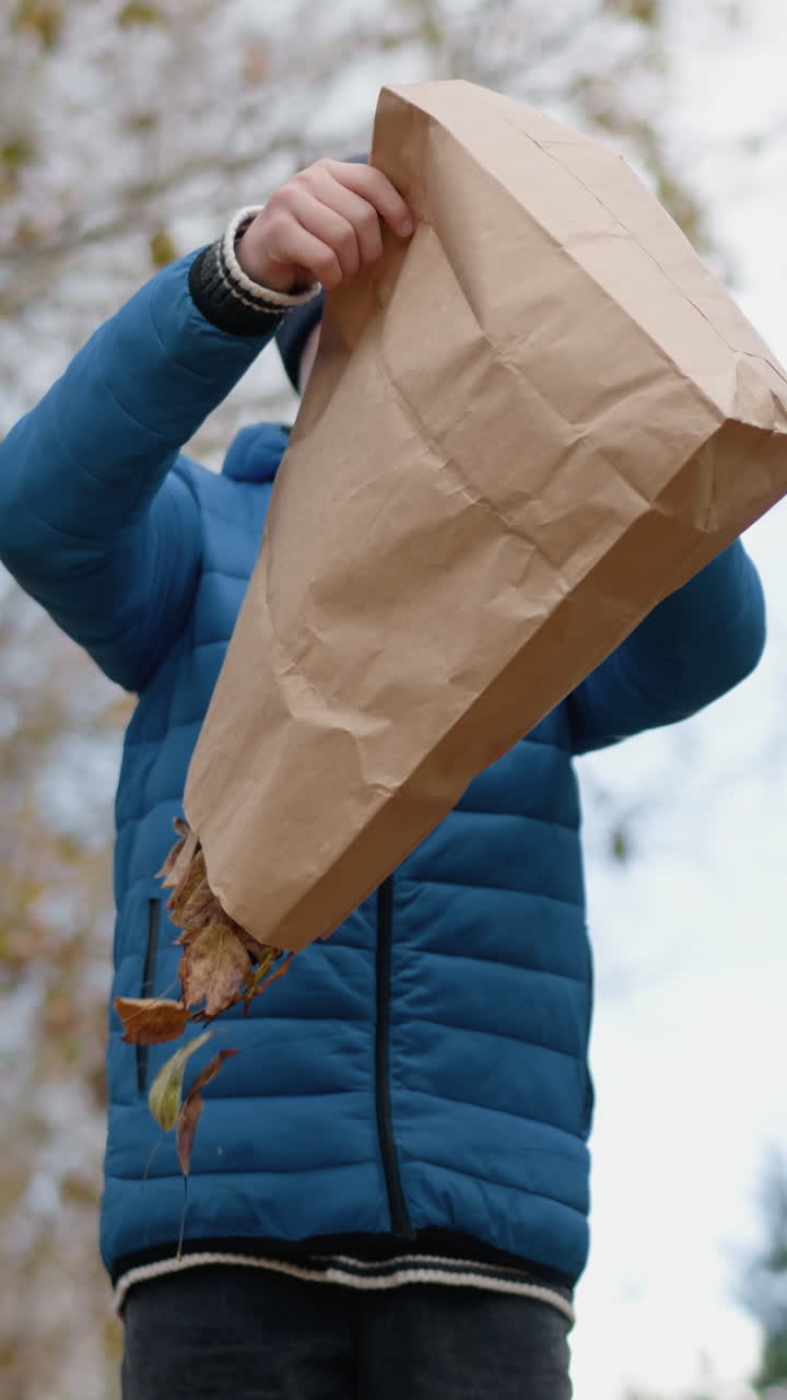 작은 소년은 마른 나무를 따라 사려 깊게 고, 그의 가방을 거꾸로 뒤집어 바닥에 건조한 잎을 뿌립니다. 다채로운 잎자루와 시골 울타리와 함께 배경에 활기찬 가을 풍경