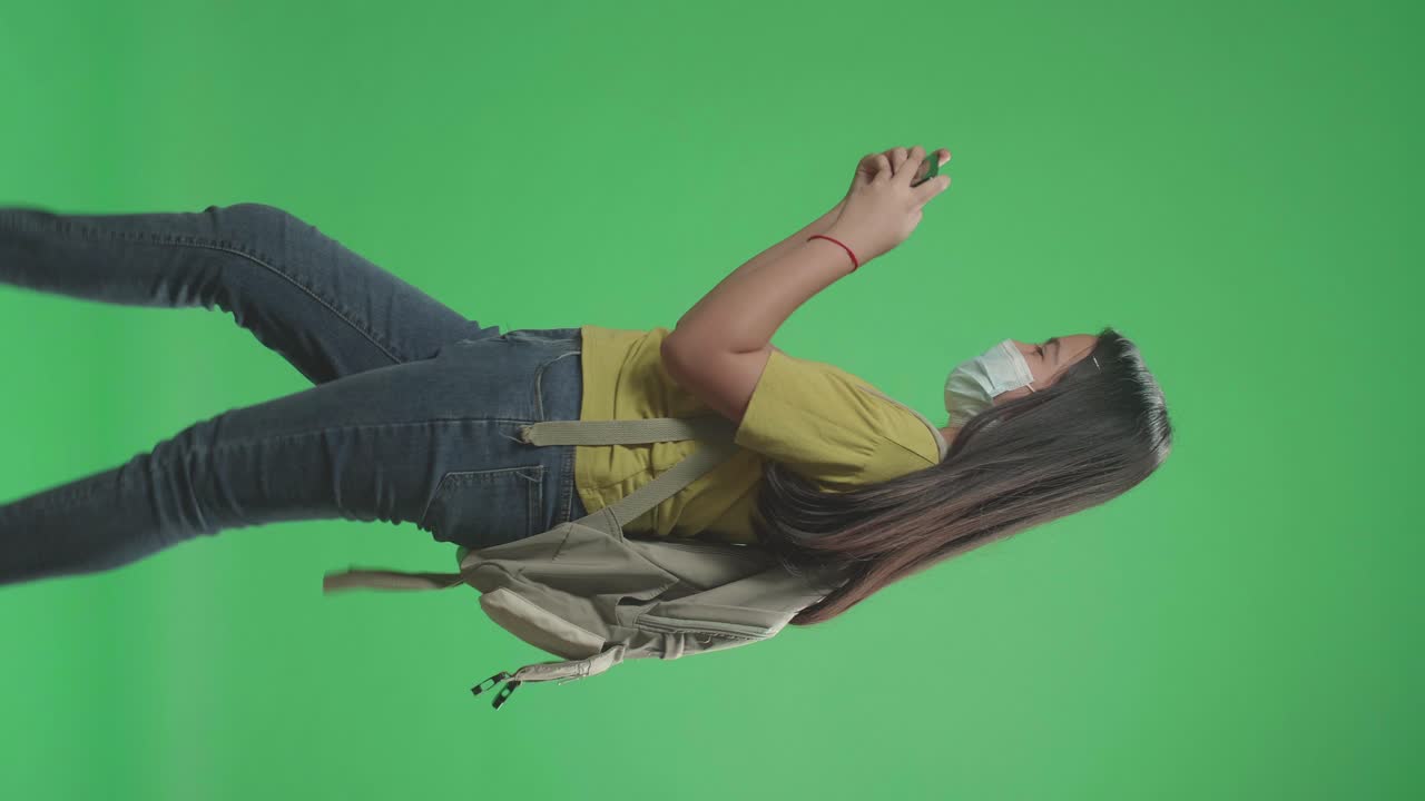 Side View Of Asian Girl Student Wearing A Mask And Playing Video Game On Mobile Phone While Walking To School On Green Screen Chroma Key