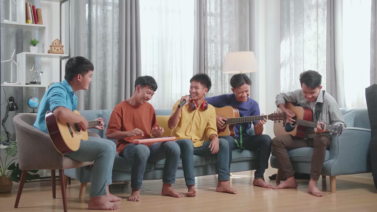 한 집 의 소파 에서 악기 를 연주 하는 동안 노래 하고 춤추는 한 친구 를 보고 웃는 젊은 아시아 친구 들 의 그룹