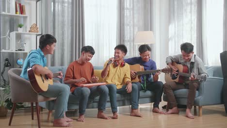 group of young asian friends laughing at one friend singing and dancing while playing musical instruments on a sofa at home