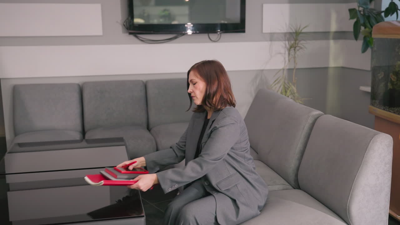 Professional woman sitting on couch in modern office, holding red notebooks, looking down at documents. Stylish office with plants and aquarium, capturing focused work atmosphere