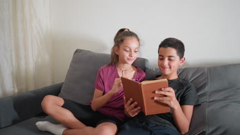 Unos amigos sentados en el sofá disfrutan estudiando juntos mientras una chica hojea un libro con una sonrisa alegre y un chico reacciona juguetonamente haciendo una expresión facial divertida durante este rato ameno.
