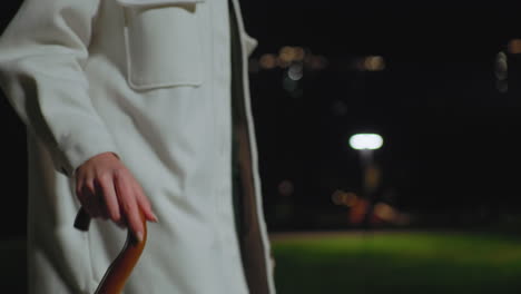 Partial view of person wearing white coat holding umbrella handle while walking outdoors at night, background featuring blurred greenery, glowing streetlights, and soft dreamy bokeh effect