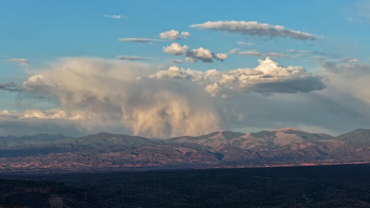 A dramatic evening timelapse capturing an isolated storm cloud releasing localized rain atop a rugged mountain range. Sweeping shadows move rapidly toward the peaks across the vast foreground valley.