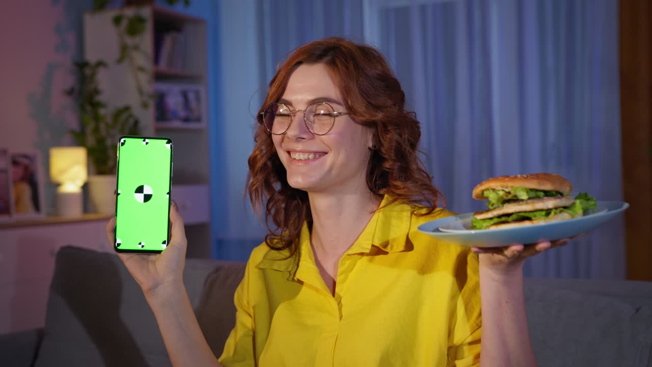 online food ordering, smiling adult girl with glasses holds phone with green chroma key on screen and burger while relaxing at home in evening