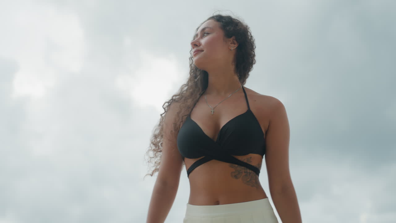 Retrato de mulher em praia de ilha sob céu dramaticamente nublado, olhar de baixo ângulo com cabelo cacheado esvoaçante pelo vento, pose de modelo confiante e tensão cinematográfica, iluminação nublada e sombria e horizonte oceânico, reflexiva