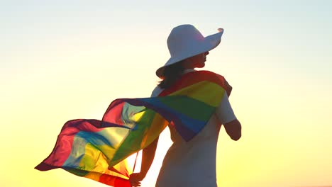 woman waving with rainbow flag and hat at sea beach at sunset