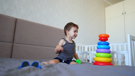 beautiful baby boy sits on a big bed playing with toys. cute peaceful toddler builds a colorful pyramid.