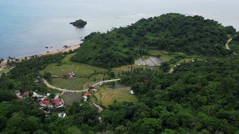 Dramatic-aerial-view-of-a-coastal,-Filipino-village-town-with-lush-forest-covered-mountains,-turquoise-ocean-waters,-and-rice-paddies