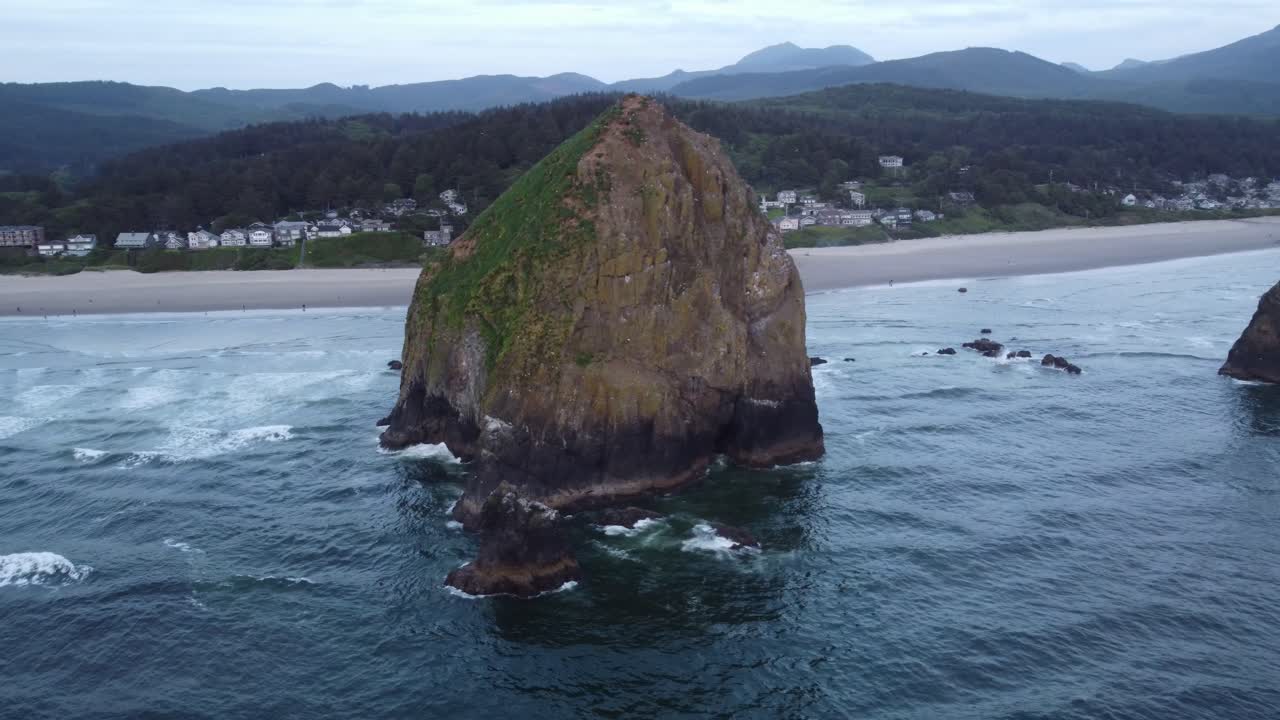 Haystack Rock in Cannon Beach, Oregon. This huge sea stack is illuminated by the sunset as waves crash onto it. Contrasted by the beach, forest, mountains, and homes in the background. 4K drone shot.