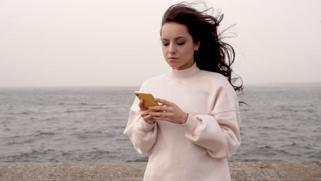 hermosa mujer usando su móvil y mirando a su alrededor, esperando a alguien junto al mar en un día ventoso