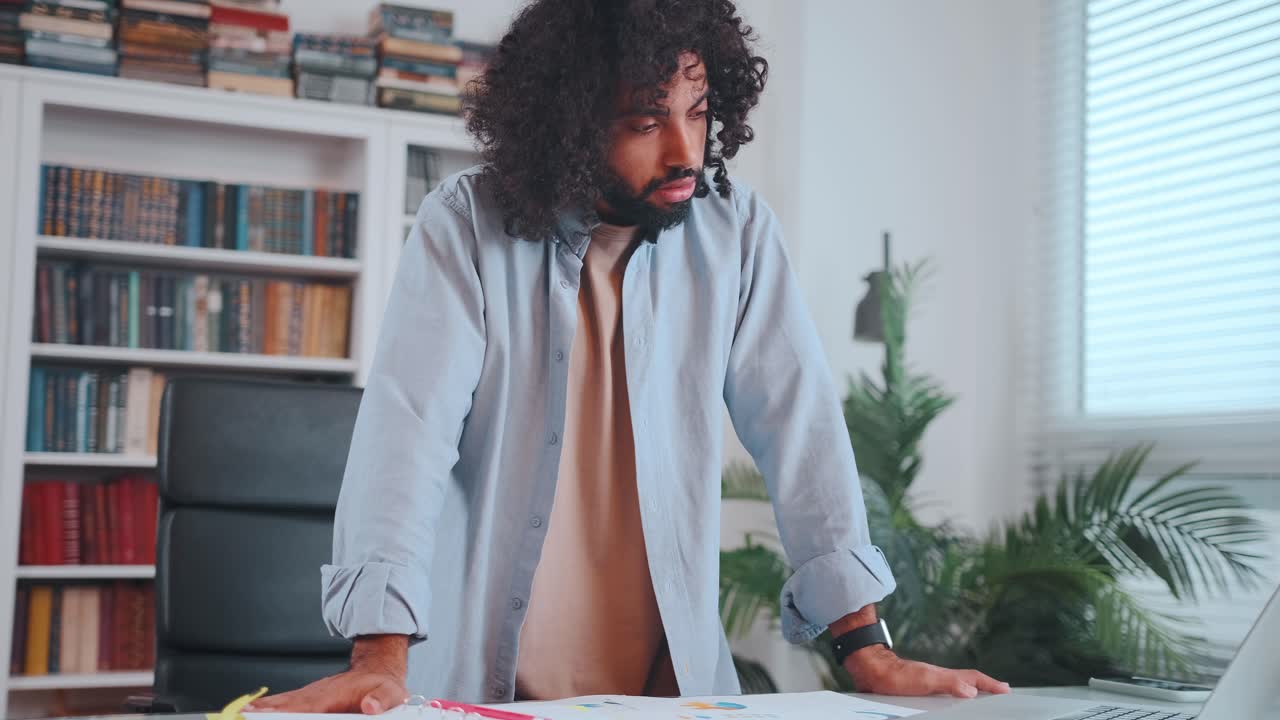 Young active arabian man works standing next to table with laptop and papers