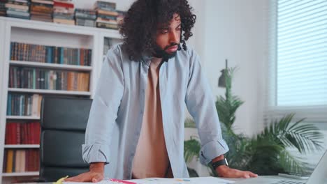 young active arabian man works standing next to table with laptop and papers