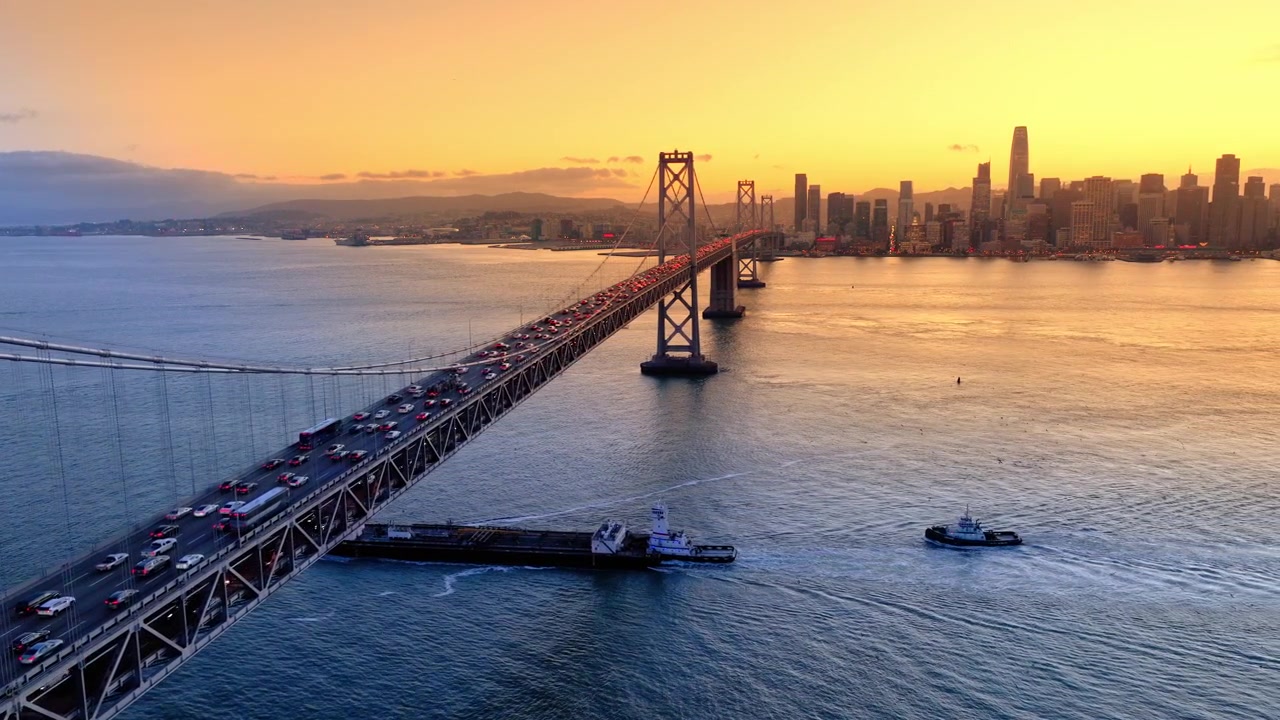 Lekter krysser under Bay Bridge ved solnedgang, San Francisco. Flyfoto av slepebåter, tett trafikk og sentrums silhuett som gløder i oransje lys.