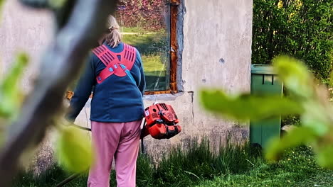 woman cutting grass in yard with brush cutter near overgrown house wall