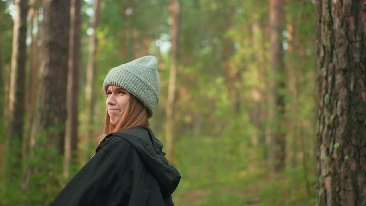 Young girl wearing gray beanie and black jacket smiling while playfully holding tent pole during camping setup in forest as sunlight filters through trees and friend stands in background