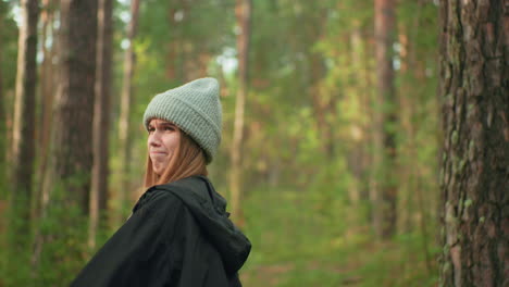 young girl wearing gray beanie and black jacket smiling while playfully holding tent pole during camping setup in forest as sunlight filters through trees and friend stands in background