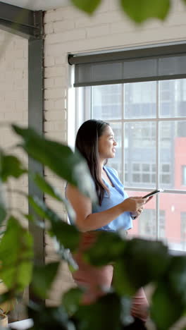 vertical video: holding tablet, businesswoman standing by window and looking outside office