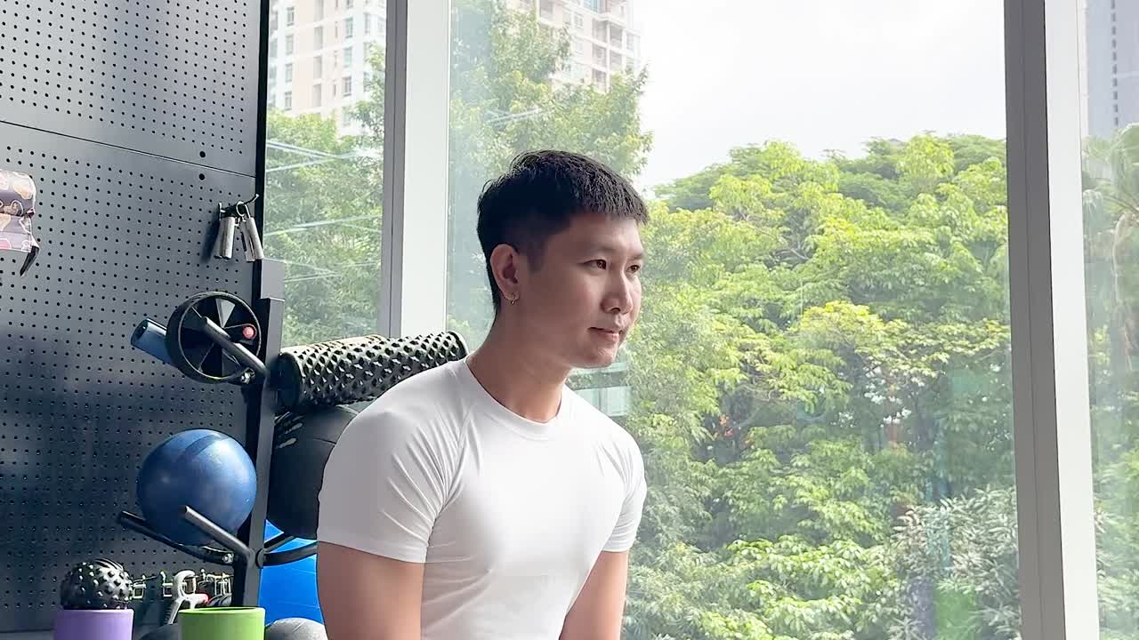 白いTシャツを着た健康的な男性が、明るいジムでケトルベルスイングをしている。