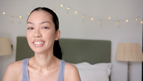 a young biracial woman with her hair pulled back smiles gently in a bedroom, with copy space