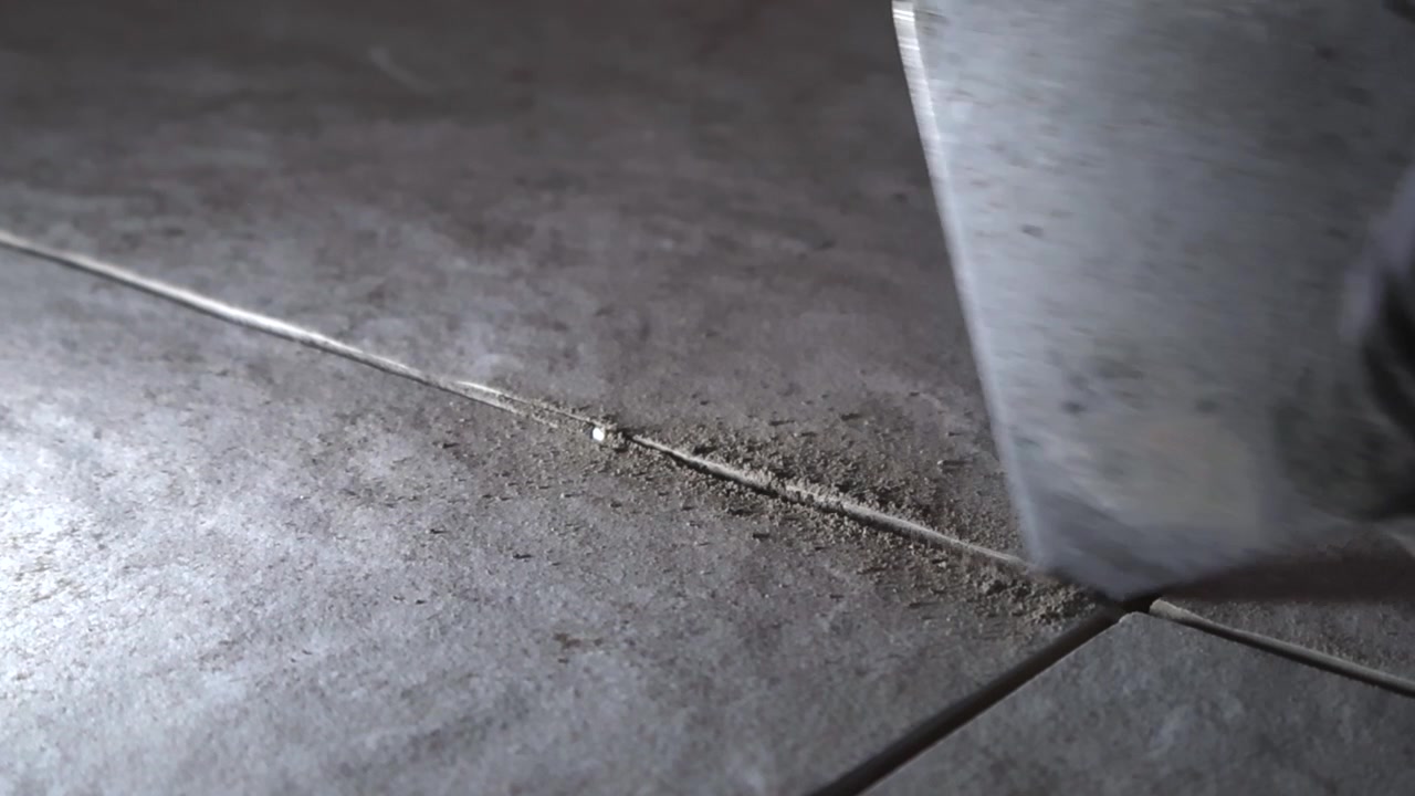 Extreme close up of a tiler wearing protective gloves and using a putty knife to clean dried mortar residue and dust from the joints between newly installed ceramic floor tiles