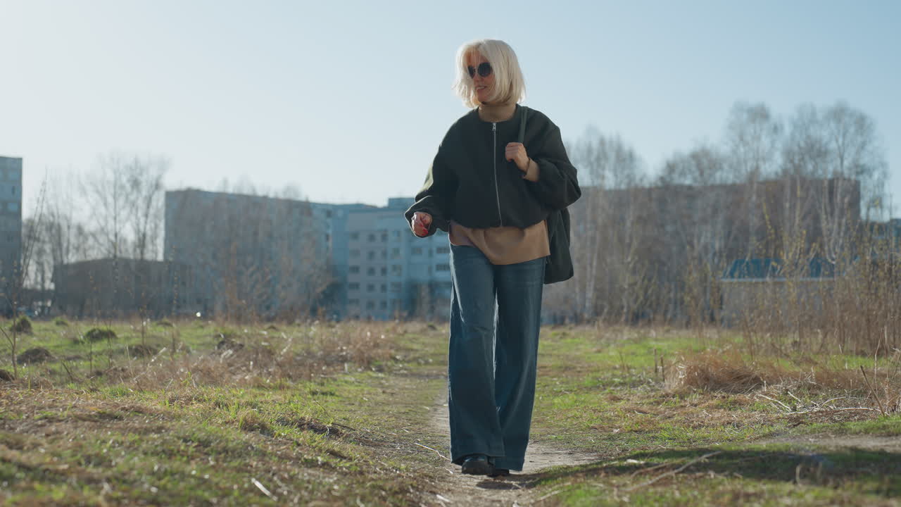 Smiling blonde woman in sunglasses wearing dark coat and jeans walking along outdoor path on sunny day with residential buildings in background, enjoying leisure stroll surrounded by nature