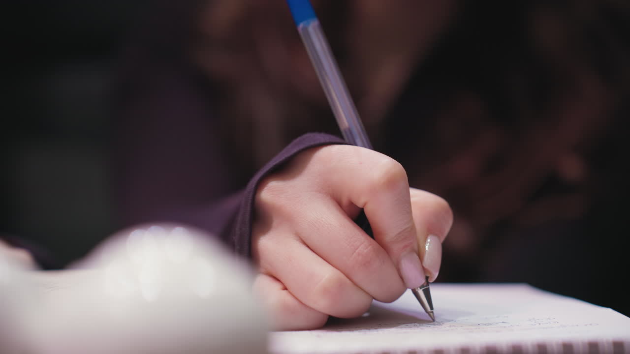 Close up of female hand holding pen while writing on notebook page. Fingernails with light polish visible, background blurred to emphasize delicate movement and attention to detail in calm atmosphere
