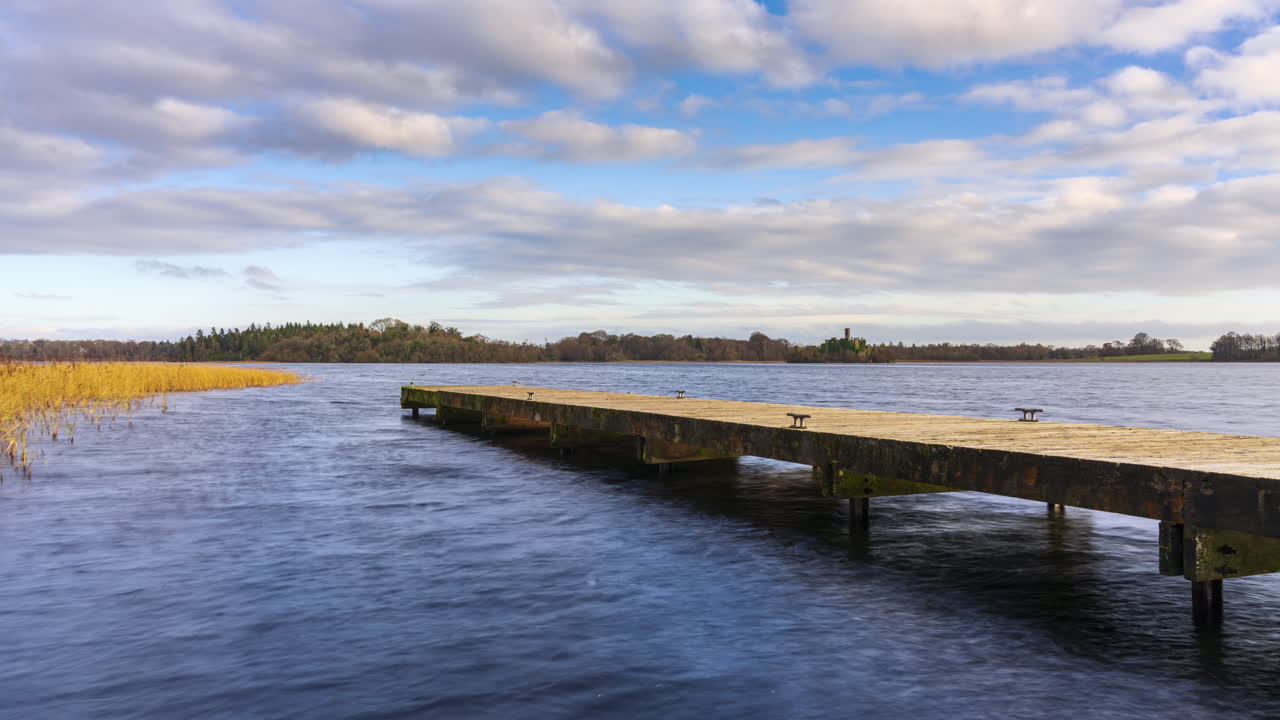 el lapso de tiempo de un muelle de lago de hormigón en primer plano y la isla de ruinas del castillo con el bosque en la distancia en un día nublado y soleado en lough key en el condado de roscommon en irlanda