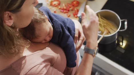 Video-of-caucasian-mother-with-newborn-baby-in-baby-carrier-cooking-in-kitchen