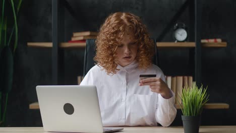 pretty red-haired girl is making online payment holding bank card using modern laptop at office table sitting and smiling. finance, shopping in internet and youth concept.