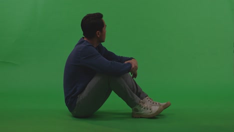 full body of distressed asian refugee male looking around then touching his head being sad due to the war while sitting on the floor on green screen background