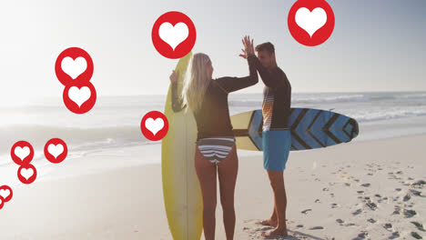 Animation-of-red-heart-love-digital-icons-over-couple-with-surfboards-high-fiving-on-beach