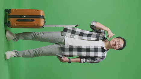 full body of asian male traveler with luggage and passport smiling and making call me gesture while standing in the green screen background studio