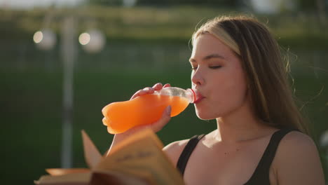 woman seated outdoors holding book in one hand and sipping juice from a bottle in the other, she continues reading, with a blurred background of greenery and lamp pole